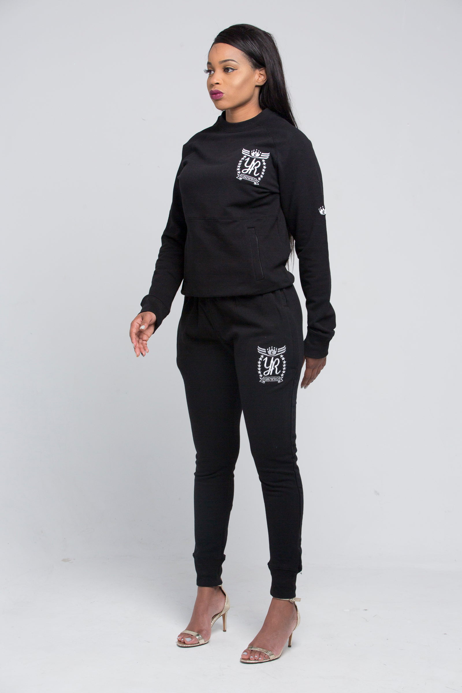 Yung'n'Rich black crew neck tracksuit shown in standing pose, featuring white logo branding on chest and thigh, slim-fit design with tapered legs and ribbed cuffs, styled with clear heels, photographed against white background