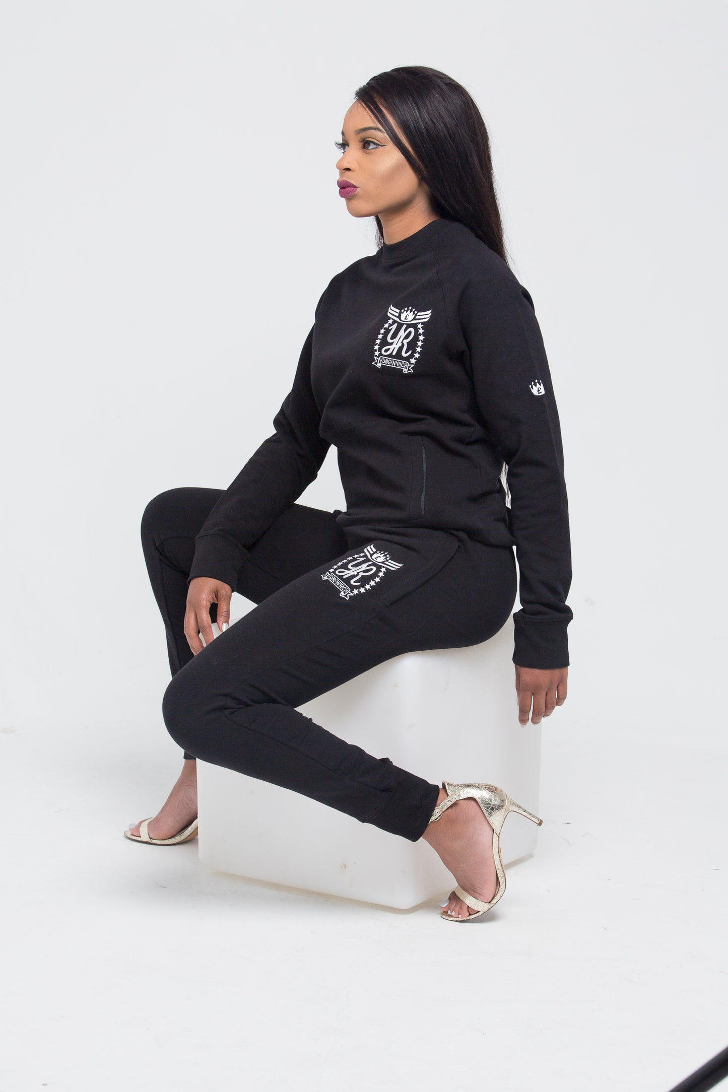 Yung'n'Rich black crew neck tracksuit modeled in seated position on white cube, showing white logo branding on chest and thigh, fitted silhouette with tapered legs, styled with clear heels, photographed against white background