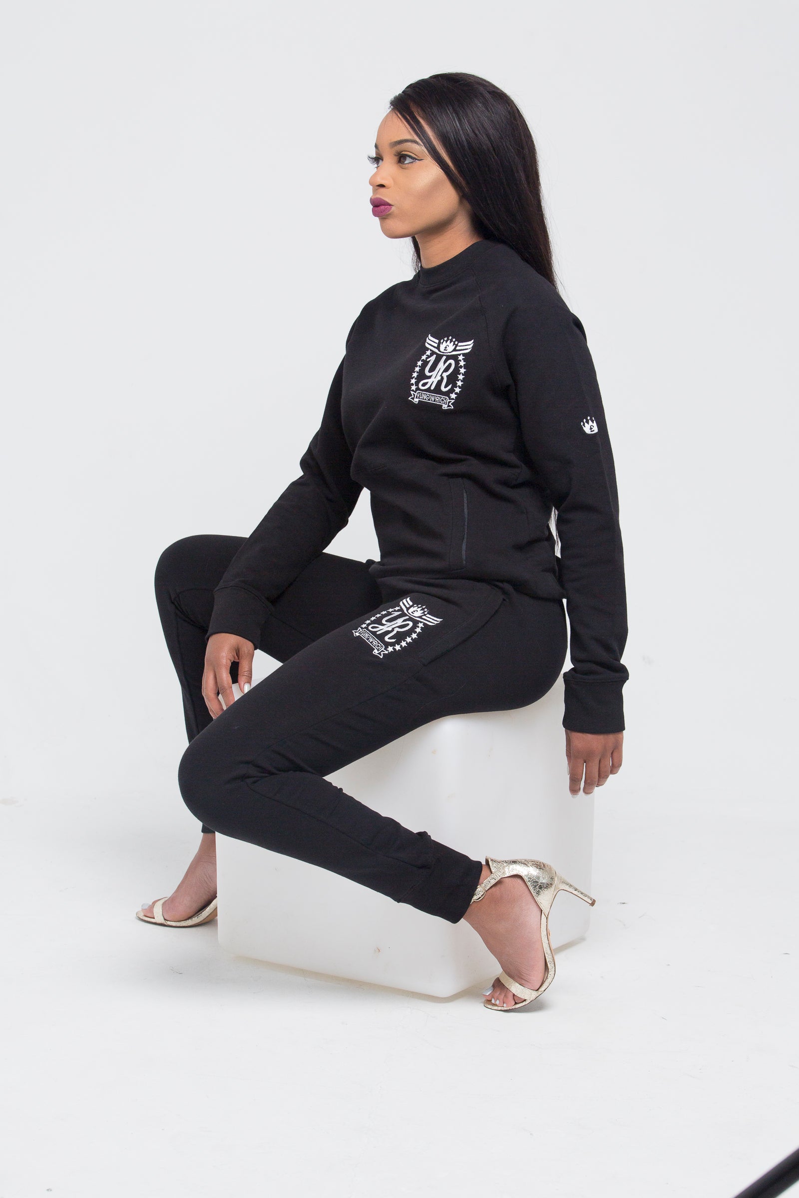 Yung'n'Rich black crew neck tracksuit modeled in seated position on white cube, showing white logo branding on chest and thigh, fitted silhouette with tapered legs, styled with clear heels, photographed against white background