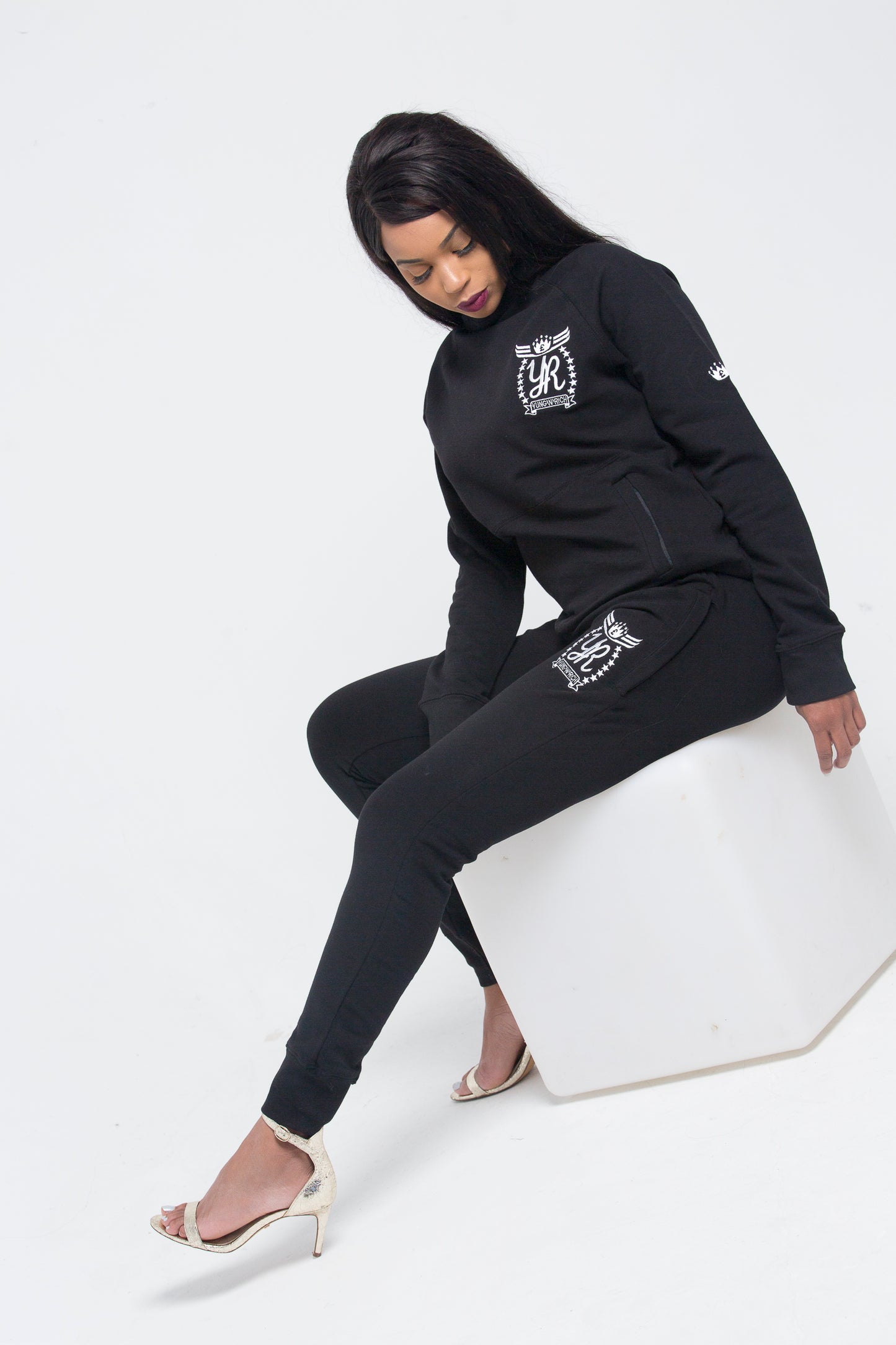 Yung'n'Rich black crew neck tracksuit shown in side-angle seated pose, displaying tapered leg fit and white logo branding on chest and thigh, styled with clear heels, photographed against white background