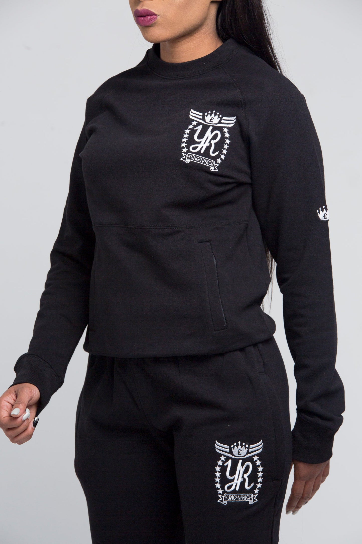 Yung'n'Rich black crew neck tracksuit detail shot, highlighting white ornate logo branding on chest and thigh, close-up view of cotton fabric texture and ribbed crew neck collar, photographed against white background