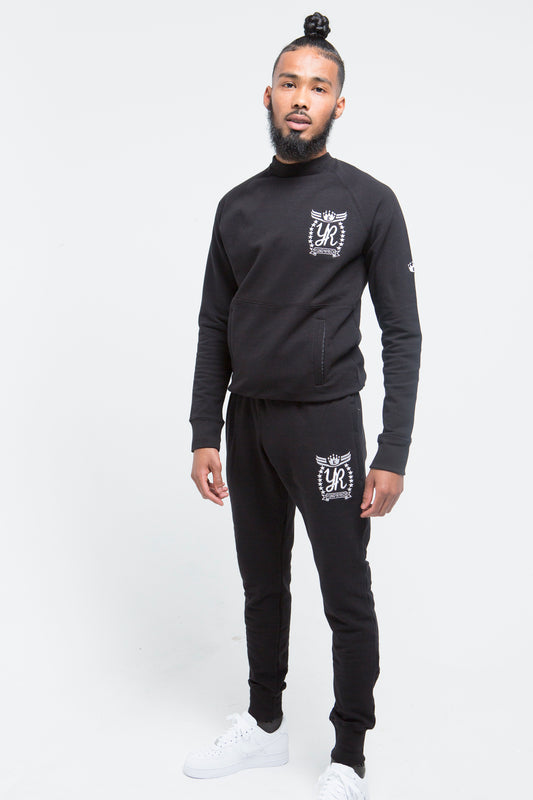 Yung'n'Rich black crew neck tracksuit on male model showing full-length view, featuring white logo branding on chest and thigh, slim-fit design with tapered legs, styled with white sneakers, photographed against white background