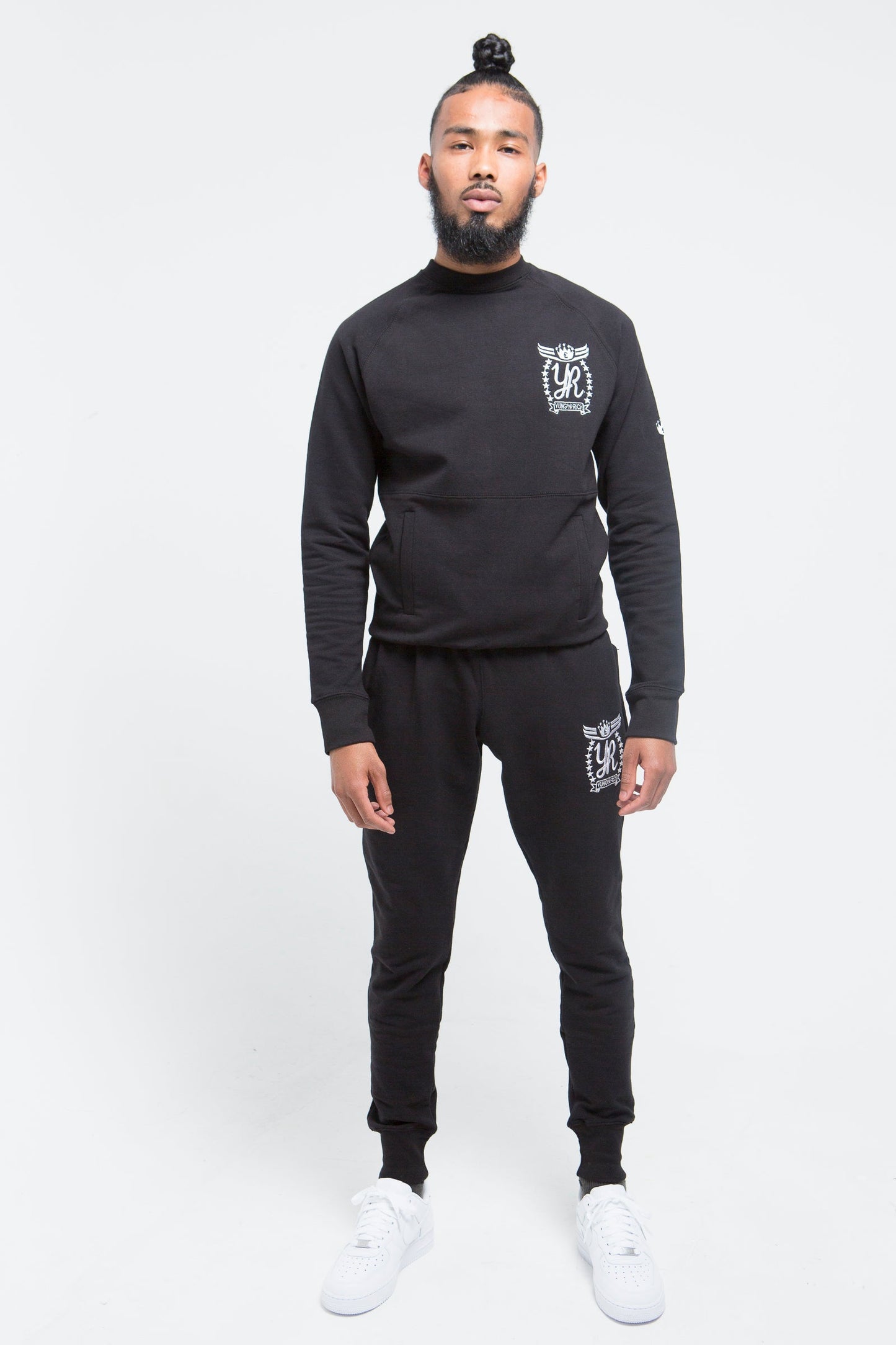 Yung'n'Rich black crew neck tracksuit front view on male model, displaying fitted silhouette with white logo placement on chest and thigh, styled with white sneakers, photographed against white background