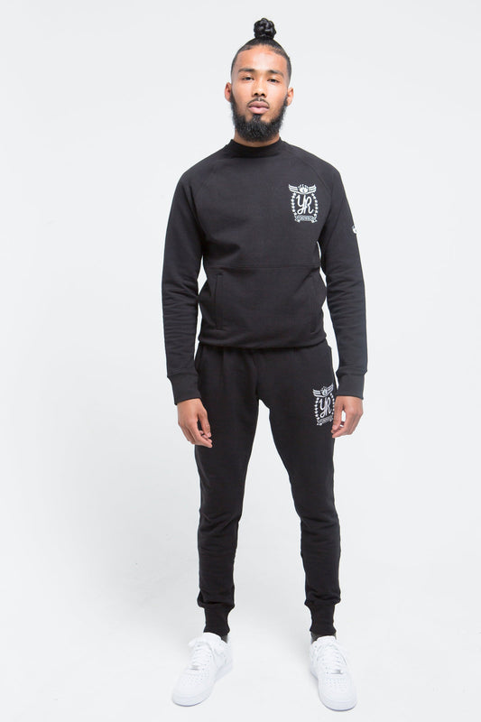 Yung'n'Rich black crew neck tracksuit front view on male model, displaying fitted silhouette with white logo placement on chest and thigh, styled with white sneakers, photographed against white background