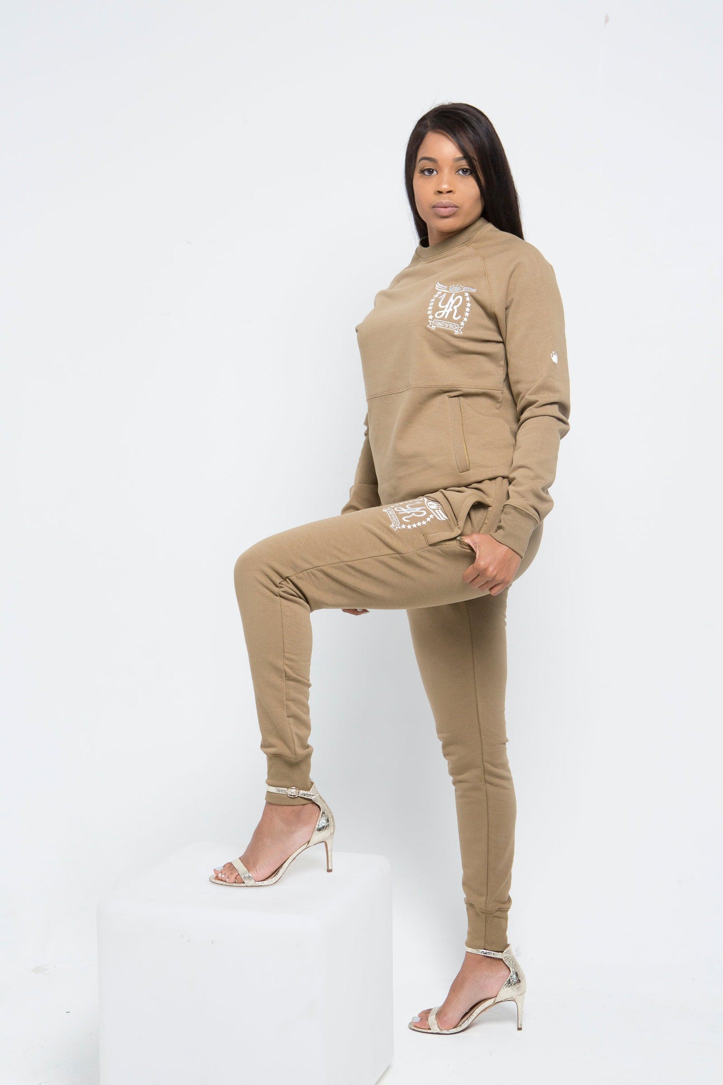 Yung'n'Rich beige crew neck tracksuit worn by a model, featuring a fitted design with ribbed collar and cuffs, tonal branding on chest and leg, shown in full-length pose against white background, styled with light colour heels