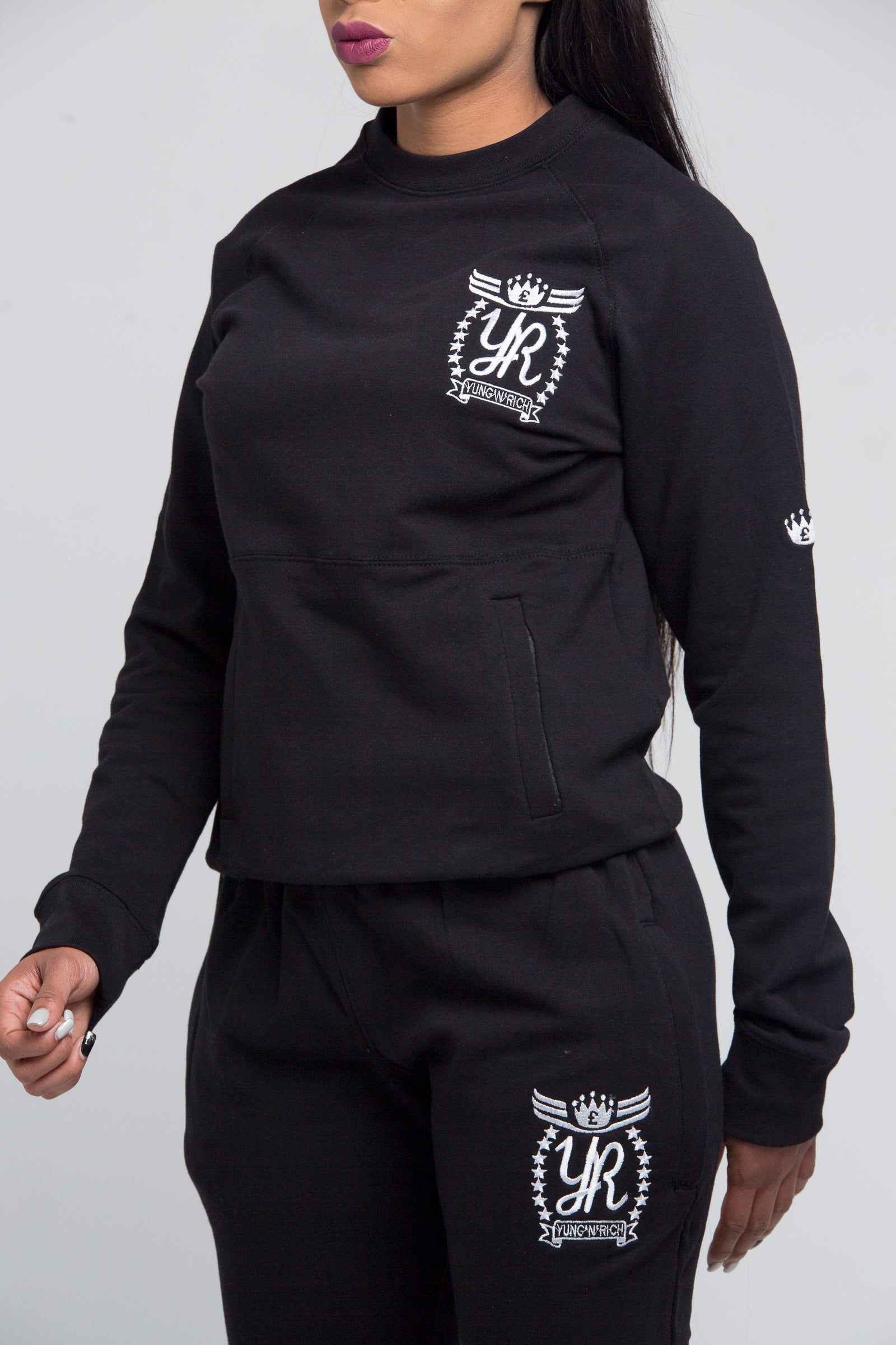 Yung'n'Rich black crew neck tracksuit detail shot, highlighting white ornate logo branding on chest and thigh, close-up view of cotton fabric texture and ribbed crew neck collar, photographed against white background